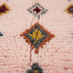 Close-up of a textured pink woven rug with a central diamond motif in mustard yellow, gray, and rust brown.