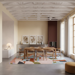 A styled dining room interior featuring a rectangular wooden dining table and six matching wooden chairs arranged on a colorful geometric area rug. The room has beige walls, a slatted wood fireplace, a white ceiling with exposed beams, and large windows with sheer curtains.