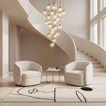A modern interior scene with two cream upholstered armchairs facing a small round side table, set on a light rug in front of a sweeping staircase and a cluster of hanging glass globe chandeliers.
