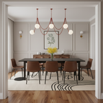 A modern dining room scene with a black rectangular dining table, six brown upholstered chairs, a red chain-link chandelier with white globe bulbs, a white rug with black curved lines, and a wood floor. The room has beige walls with white molding, wall sconces, and a framed botanical print.