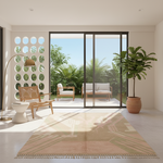 A large rectangular area rug with fringe, in beige, blush, and muted green tones, styled in a bright modern living room with sliding glass doors leading to a garden patio.