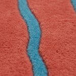 Close-up of a textured rug or fabric with a coral-red field and a wavy vertical blue stripe running through the center.