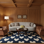 A styled living room scene with a light gray sofa centered against wood-paneled walls, flanked by two brown armchairs. A round dark wood coffee table sits on a navy blue area rug with large cream hearts.