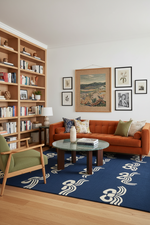 A styled living room interior featuring a terracotta orange tufted sofa, a green armchair, a blue rug with white wave patterns, a wooden bookshelf, and a framed landscape print on the wall.