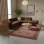A styled living room scene featuring a brown sectional sofa, a round coffee table, and a large red and beige patterned area rug on a light gray floor. The room includes a plant, wall art, a floor lamp, and a wooden chair.