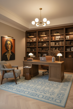 A styled home office interior featuring a large wooden writing desk and matching bookshelves, with a blue patterned area rug, lit pendant ceiling chandelier, and a framed portrait on the wall.