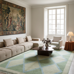 A styled living room featuring a large beige sectional sofa, a light green and cream geometric area rug, a dark wood coffee table, a white armchair, and a large classical tapestry on the wall.