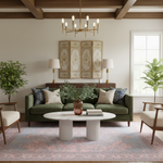 A styled living room scene featuring an olive green sofa with patterned throw pillows, a round white coffee table, a pink patterned rug, and a gold chandelier in a room with exposed wooden ceiling beams.