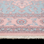 A close-up of a patterned area rug showing its light pink field, pale blue ornamental motifs, and cream-colored fringe along the bottom edge.