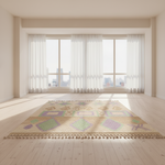 A rectangular area rug with a geometric pattern in beige, green, purple, and orange, laid out on a light wood floor in a bright, modern room with large windows and white curtains.