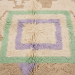 A close-up of a shaggy wool rug with concentric square patterns in cream, light green, lavender purple, and beige.
