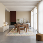 A modern dining room scene with a rectangular wooden dining table and matching chairs placed on a light patterned rug. The room has beige walls, white ceiling beams, sheer curtains, a sideboard with framed art, a dark ribbed wall panel, and a round brown ottoman in the foreground.