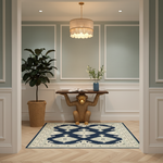 A rectangular area rug with a navy blue and cream geometric pattern is displayed on a light wood herringbone floor in a styled entryway. The room features a console table with a monkey sculpture, a potted plant, and a hanging chandelier.