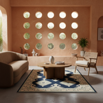 A styled interior scene with a round wooden coffee table centered on a navy and beige patterned area rug. A beige sofa sits on the left, a mid-century style chair with a cane back is on the right, and the room has warm peach stucco walls with circular cutouts and a potted plant.