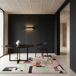 A modern interior scene with a round black dining table, a black chair, a white vase, and a colorful geometric area rug on a light stone floor. The room has a dark wood-paneled wall, a black wall sconce, and a textured ceiling.