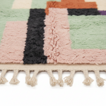 Close-up of a textured geometric area rug with a chunky shag pile and fringe tassel, featuring blocks of dusty pink, sage green, black, orange, and cream on a white background.