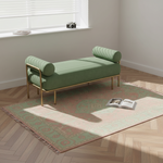 A green striped chaise lounge with a gold metal frame sits on a patterned rug in a bright room with wood herringbone flooring and a window.