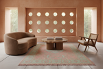 A styled interior scene with a curved upholstered sofa on the left, a mid-century wooden armchair on the right, and a round coffee table centered on a patterned rug. The room has warm terracotta walls, circular window cutouts, and large windows looking out to greenery.
