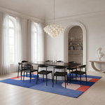 A dining room scene with a black rectangular dining table and six black chairs placed on a large blue, pink, and coral area rug. The room has white walls, tall windows with white curtains, a white pendant chandelier, and an arched built-in niche in the background.
