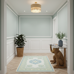 A styled entryway or hallway with pale green walls, white wainscoting, a hanging crystal chandelier, a patterned area rug, a potted plant, and a glass-top console table with a carved wooden seat.