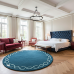 A styled bedroom interior featuring a large round teal blue rug with a white decorative border, placed on a light wood herringbone floor. The room includes a bed with a tufted navy headboard, white bedding, a red sofa, a red armchair, and a black chandelier.
