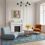 A styled living room interior featuring a light blue ribbed lounge chair, an orange armchair, a round white coffee table with yellow flowers, a colorful blue patterned area rug, and a white fireplace with a framed abstract painting.