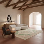 A styled interior room with a cream curved sofa, dark gray armchair, wooden side table, and a large patterned area rug on a terracotta floor. The room has exposed dark wood ceiling beams, arched windows, and a minimalist black-and-white wall art piece.