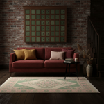 A styled living room scene featuring a rectangular area rug with a traditional geometric pattern in sage green, beige, and dusty pink. The rug is placed on a dark wood floor in front of a red velvet sofa, with a brick wall and a wooden decorative screen behind it.