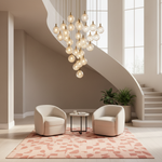 A modern multi-light cluster chandelier with glowing glass globe bulbs hangs in a bright, contemporary stairwell lobby. Two cream-colored armchairs and a small round side table sit on a pink patterned rug, with a staircase and large windows in the background.