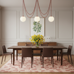 A styled dining room scene with a dark wood rectangular table, four brown chairs, a pink patterned rug, pendant lights, and a vase of yellow flowers against a light paneled wall.