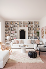 A bright, modern living room featuring a large white sectional sofa with blush and gray pillows, set in front of a massive white built-in bookshelf. The room has pink-tiled floors, a chevron-patterned rug, a black and white pouf, and a wicker armchair.
