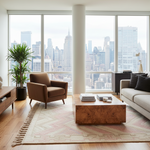 A modern living room with a brown armchair, a beige sofa, a wooden coffee table, and a patterned rug, all set against a large window with a city skyline view.