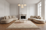 A modern living room featuring two curved beige sectional sofas facing a round coffee table on a patterned chevron rug. The room has white paneled walls, a white marble fireplace, herringbone wood floors, and three hanging pendant lights.