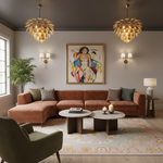 A styled living room featuring a terracotta-colored sectional sofa with matching throw pillows, two round marble-top coffee tables, a patterned area rug, a green armchair in the foreground, and two distinctive leaf-shaped chandeliers hanging from a dark ceiling. A colorful painting is mounted on the wall above the sofa.