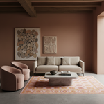 A styled living room interior featuring a light beige sofa, two blush pink armchairs, a stone-topped coffee table, a patterned pink and cream rug, and warm terracotta walls with framed art.