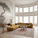 A modern living room featuring a mustard yellow sectional sofa with dark gray accent pillows, set on a pink and blue patterned area rug. The room has a large window bay, a fireplace with a black-and-white landscape mural on the wall, and a round wooden coffee table.