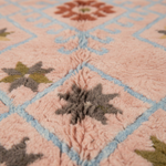 Close-up of a textured, loop-pile rug with a dusty pink base, light blue diamond outlines, and gray and olive green starburst medallions.