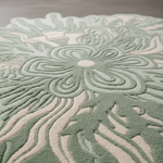 A close-up of a textured area rug with a raised floral pattern in sage green and cream, showing the detailed pile and depth of the design.