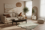 A styled living room scene with a cream sofa, dark wood coffee table, pale green patterned rug, woven pouf, mirror, plant, pendant light, and abstract wall art in a warm neutral interior.