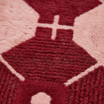 Close-up of a textured woven rug or textile in deep burgundy red and pale blush pink, showing a geometric pattern with a central cross motif.