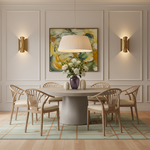A styled dining room scene featuring a round gray concrete table surrounded by six light wood spindle-back chairs with cream cushions. A large pleated white drum pendant light hangs above the table, which is decorated with a purple vase of white flowers and two textured white side tables.