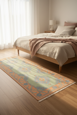 A rectangular area rug with a traditional medallion pattern in sage green, orange, and blue, styled on a light wood floor in a bright, modern bedroom next to a wooden bed.
