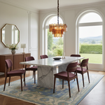 A modern dining room scene featuring a rectangular white marble dining table with a white marble pedestal base, surrounded by six mid-century modern chairs with burgundy upholstery and dark wood frames. The set is styled on a blue and beige patterned area rug under a glass chandelier, with large arched windows showing a green garden view.