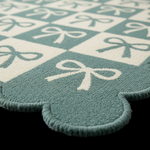 A close-up of a plush textured rug with a scalloped edge, featuring a mint green and cream checkerboard pattern with white bow motifs.