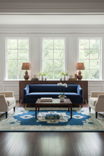 A styled living room featuring a deep blue three-seater sofa as the central product, set on a blue and beige patterned area rug with a dark wood coffee table, flanked by two cream armchairs. The room has white walls with decorative molding, three large windows showing green trees outside, and a dark wood console table with matching lamps.