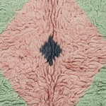 A close-up of a textured, looped fabric or rug with a diamond-shaped dusty pink center, a small dark charcoal diamond motif in the middle, and a sage green border.