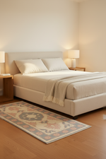 A modern upholstered bed in a warm neutral bedroom setting, featuring a cream-colored bed frame, matching bedding, a beige throw blanket, a patterned area rug, and wooden nightstands with lit lamps.