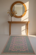 A rectangular area rug with a pink and green traditional medallion pattern and fringe, styled on a white floor in front of a light wood console table and a round wooden mirror.