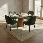 A modern dining set in a bright, elegant room featuring a square table with a ribbed gold pedestal base and a white marble top, surrounded by four dark green velvet chairs. A chess set is placed on the table, and the furniture is styled on a patterned rug with a wood parquet floor.