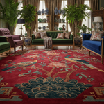 A large traditional oriental area rug with a red background, blue and green wave motif, and bird-and-floral patterns, shown in a styled living room with green, blue, and purple sofas, plants, and shelving.